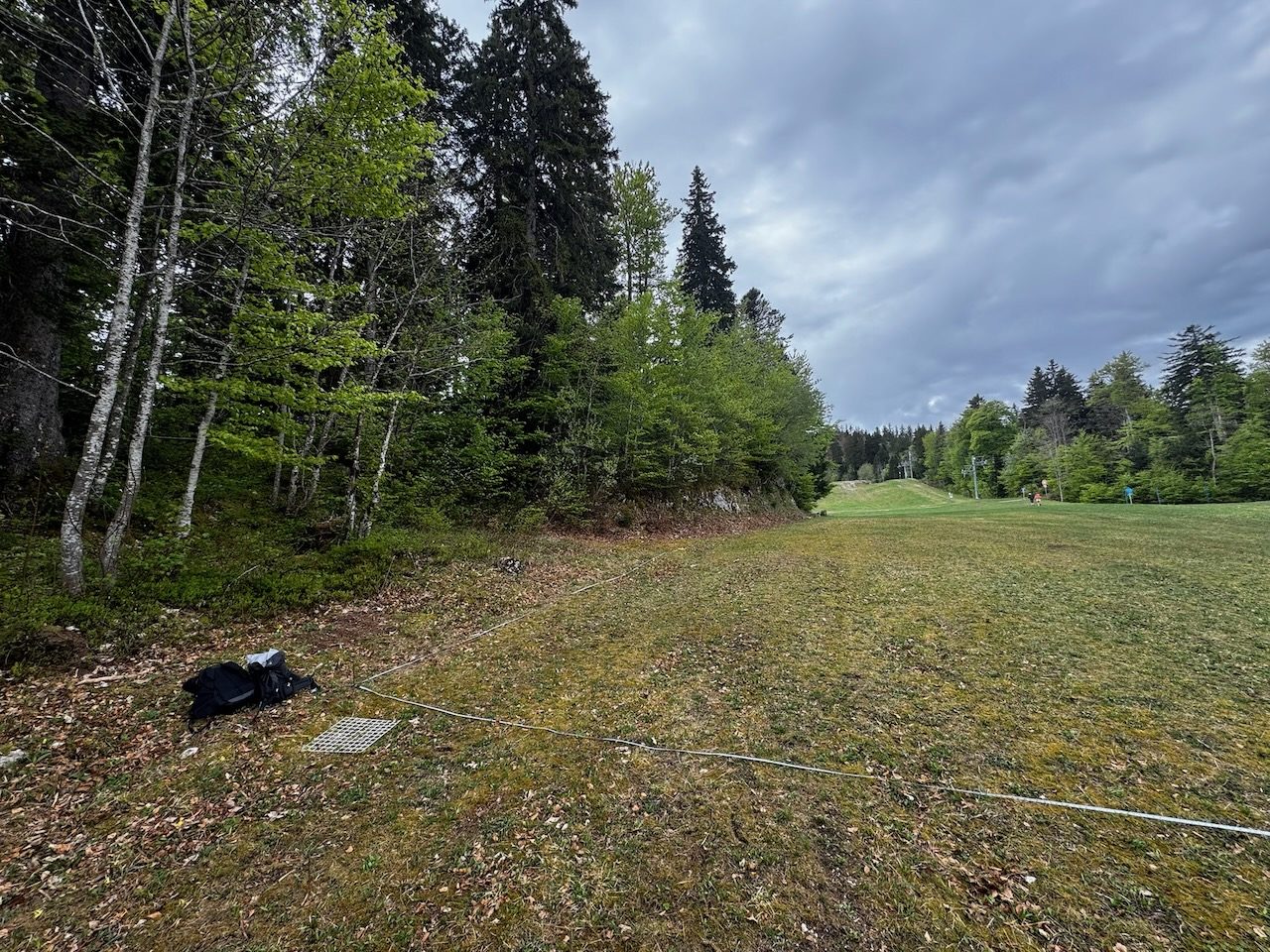 The piste edge at Mijoux from ground level: mixed forest along the left, mown grass to the right, a measuring tape running out from under the trees across the boundary, a quadrat frame and a rucksack on the ground, and a ski lift in the distance.